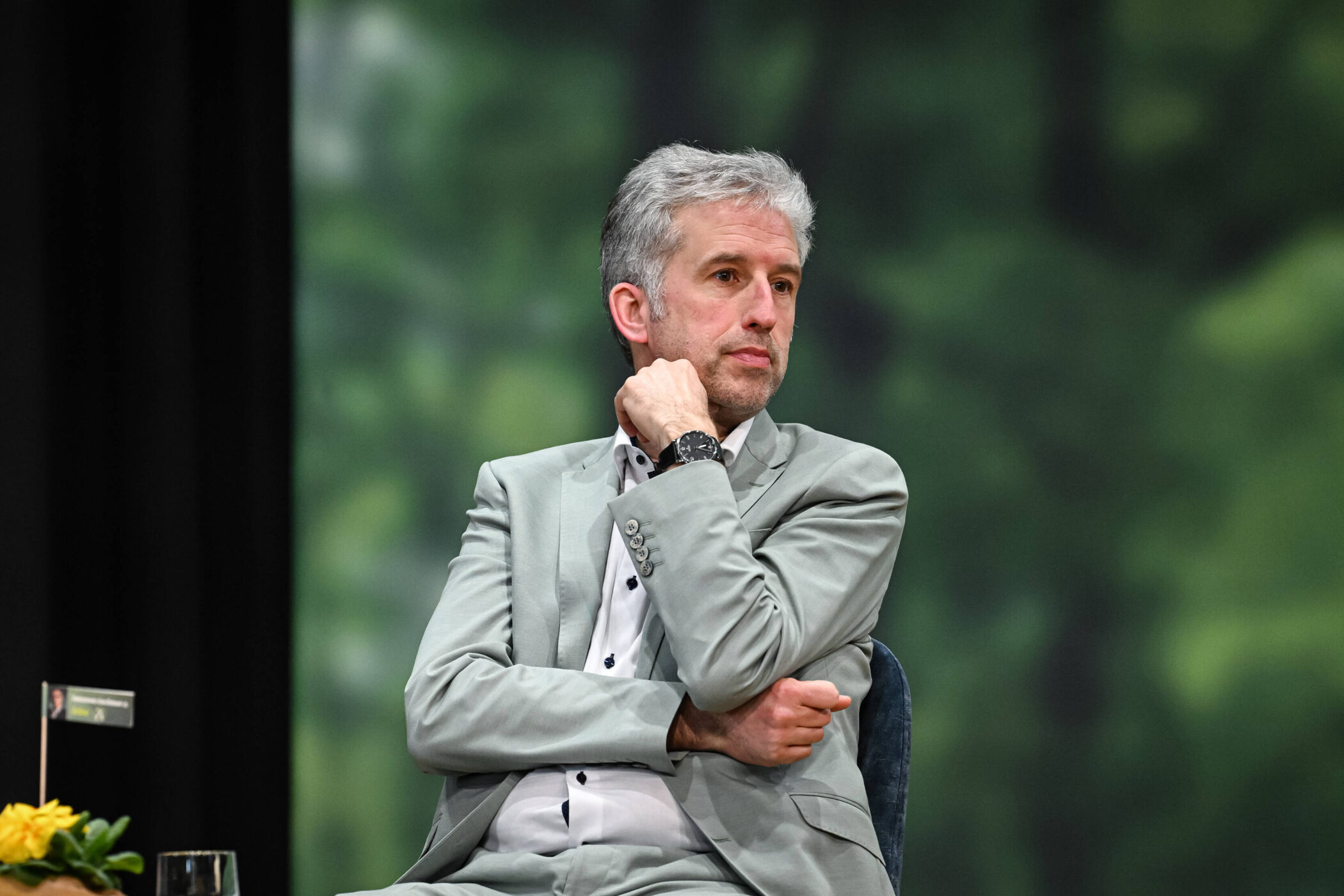 Mann in grauem Anzug sitzt, stützt Kinn auf Hand. Blumen im Vordergrund.