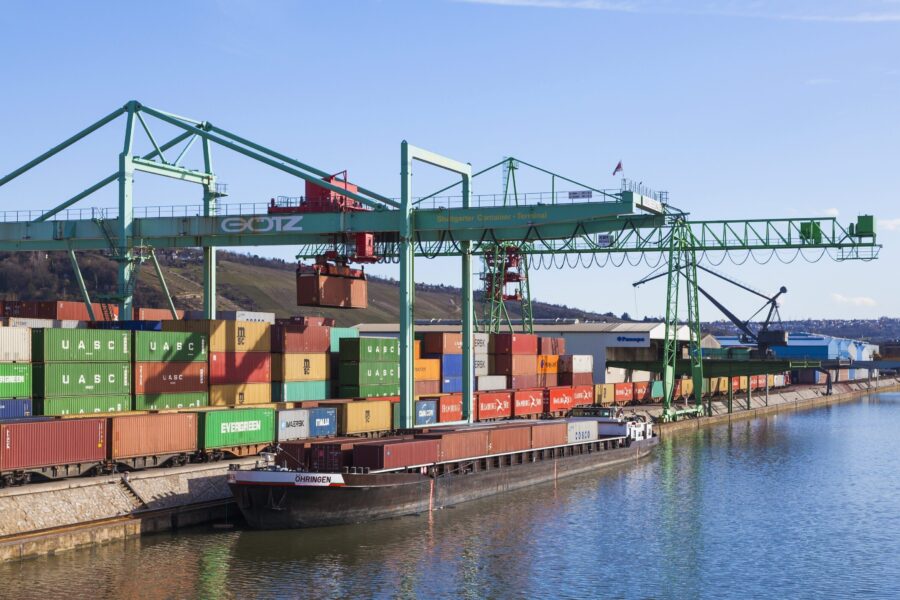 Hafen mit Containern, Kranen und einem Schiff am Ufer unter blauem Himmel.