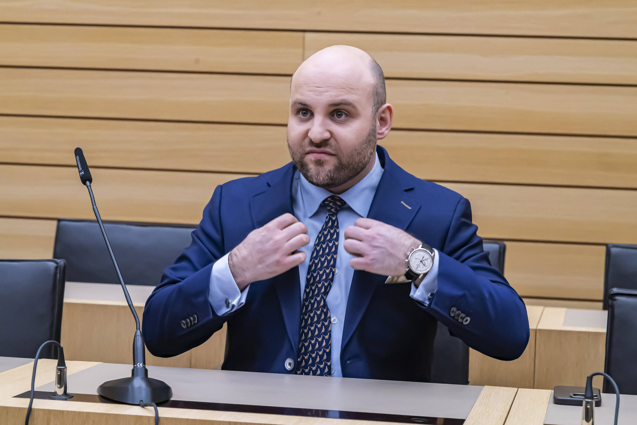 Person in Anzug sitzt an Tisch, berührt Krawatte; Mikrofon steht vor ihm.