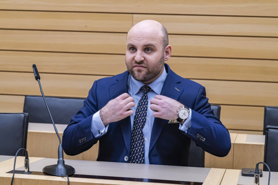 Person in Anzug sitzt an Tisch, berührt Krawatte; Mikrofon steht vor ihm.