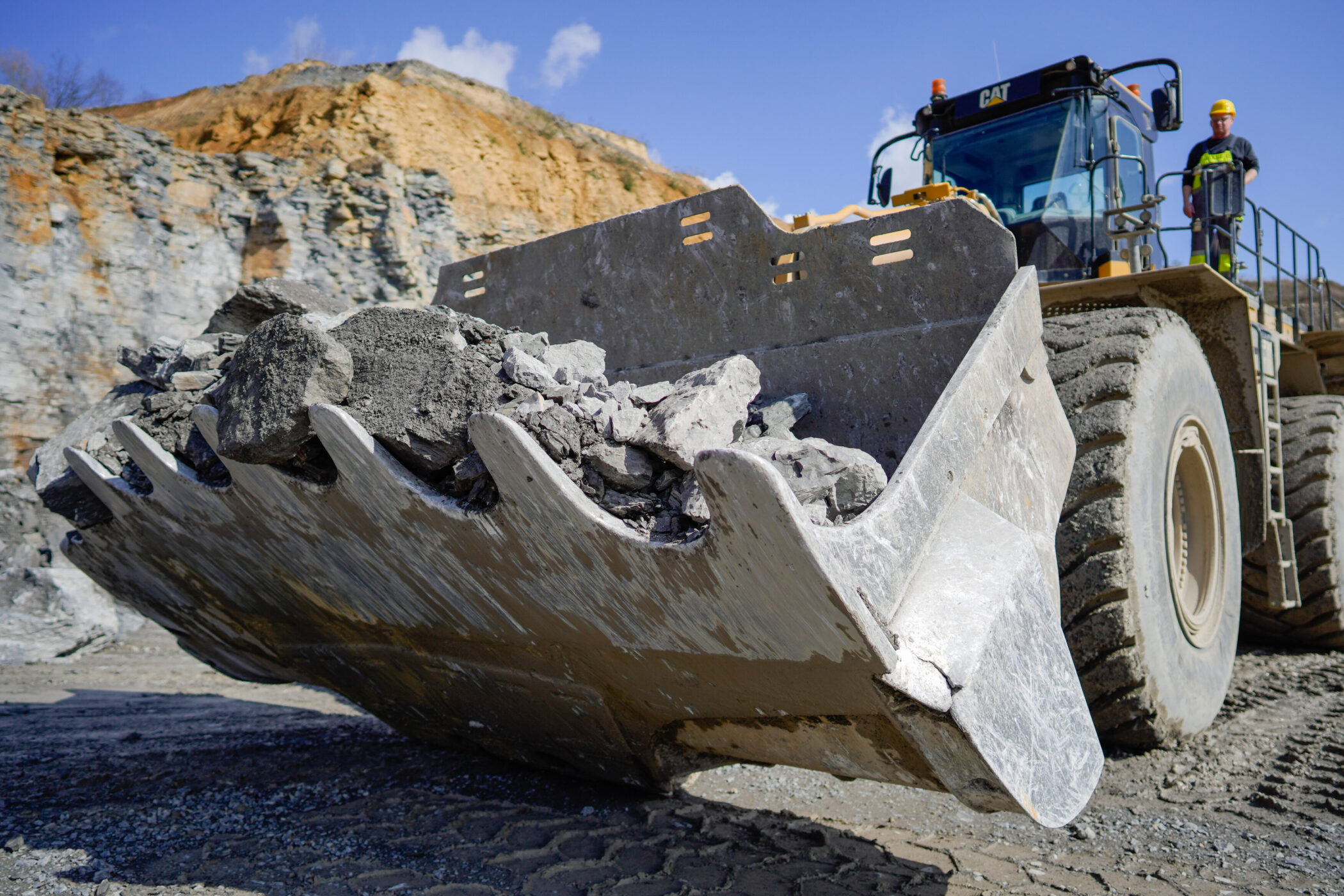 Großer Bagger mit voller Schaufel, zwei Arbeiter stehen daneben im Steinbruch.