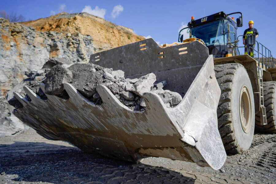 Großer Bagger mit voller Schaufel, zwei Arbeiter stehen daneben im Steinbruch.
