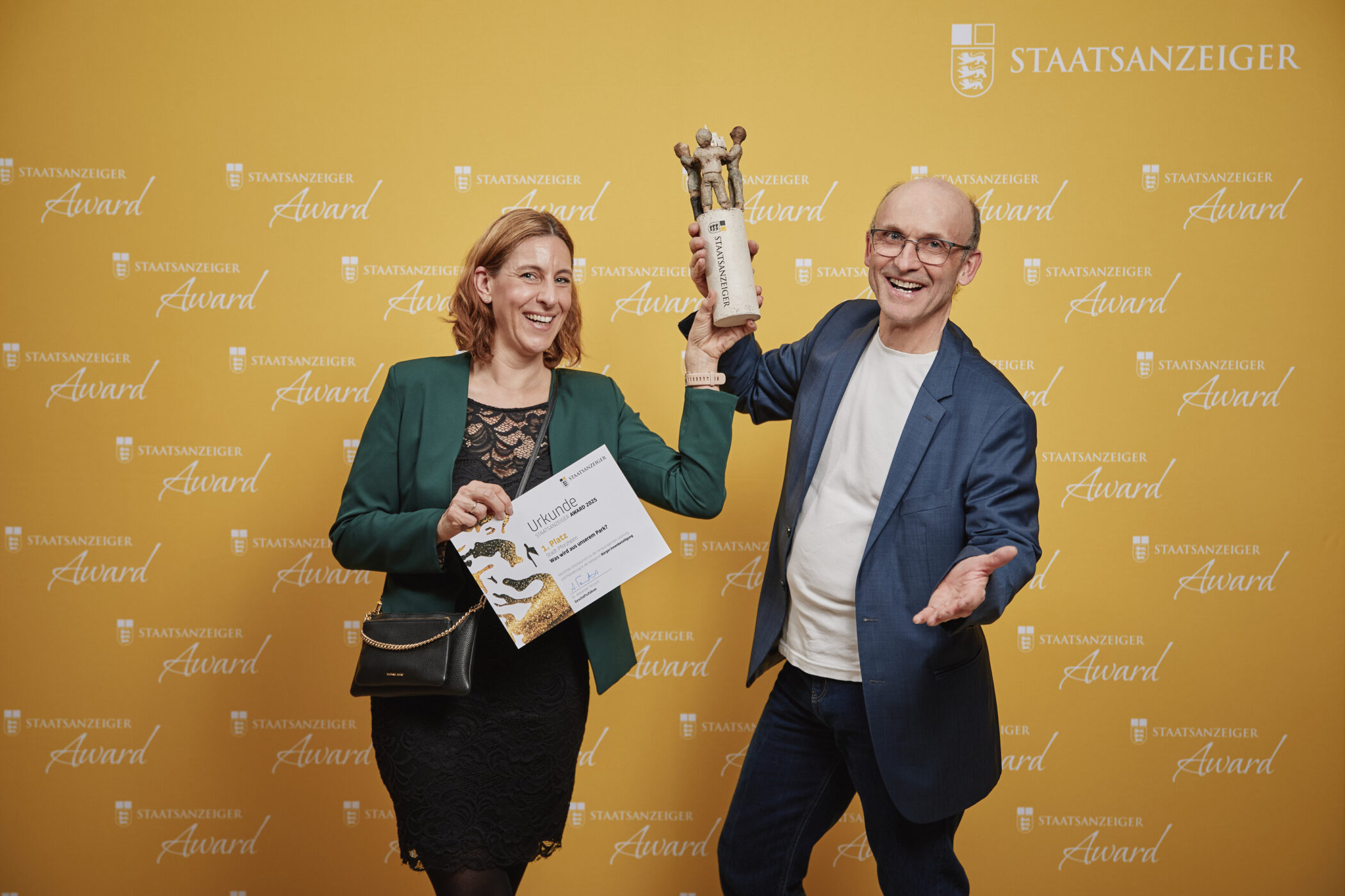 Zwei Personen halten eine Urkunde und eine Trophäe vor gelbem Hintergrund.