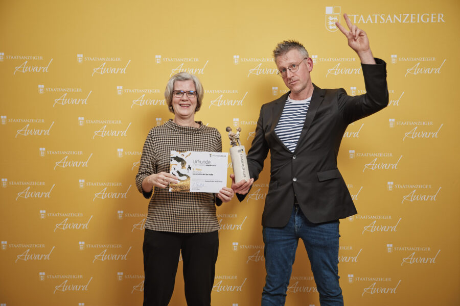 Zwei Personen vor gelber „Staatsanzeiger Award“-Wand, halten Urkunde und Trophäe.
