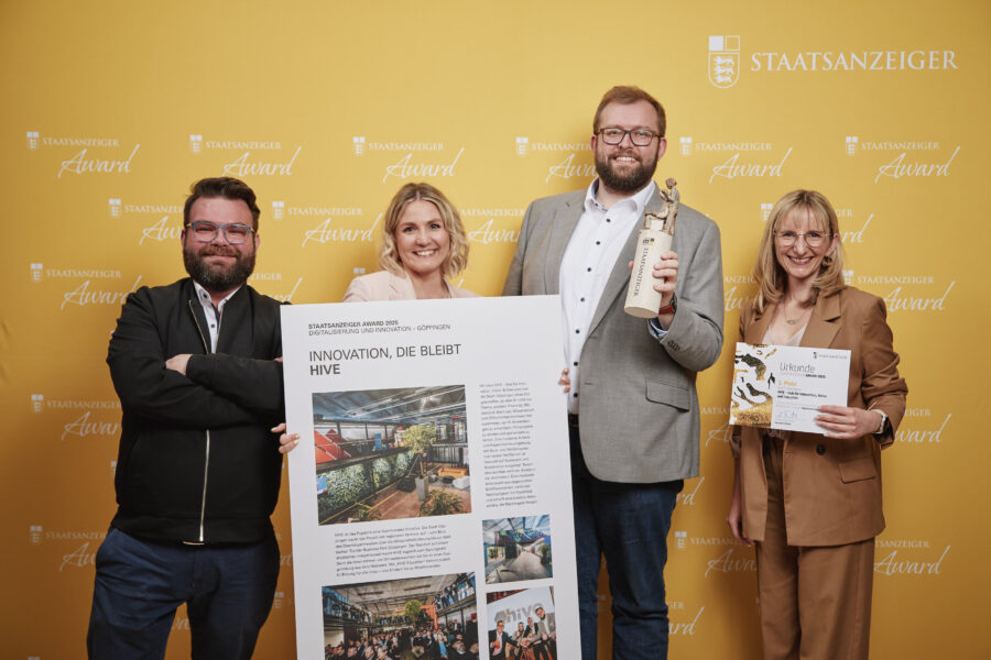 Vier Personen vor gelbem Hintergrund mit Preis und Urkunde, Text: „Staatsanzeiger Award“.