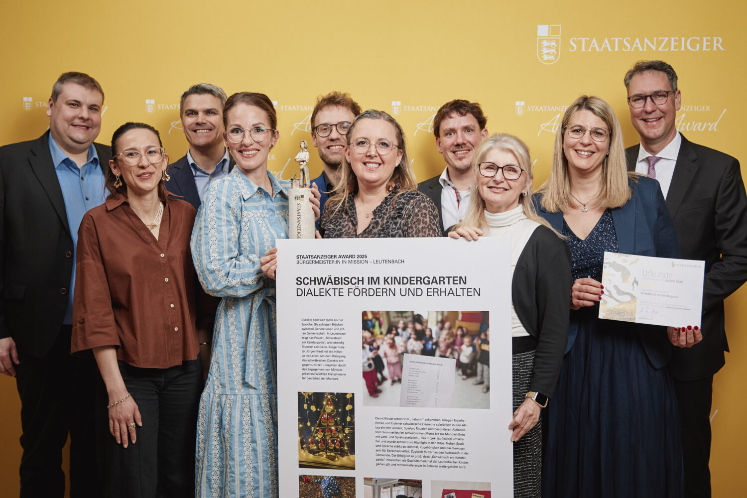 Zehn Personen mit Urkunde und Plakat vor gelbem Hintergrund. Einer hält eine Trophäe.