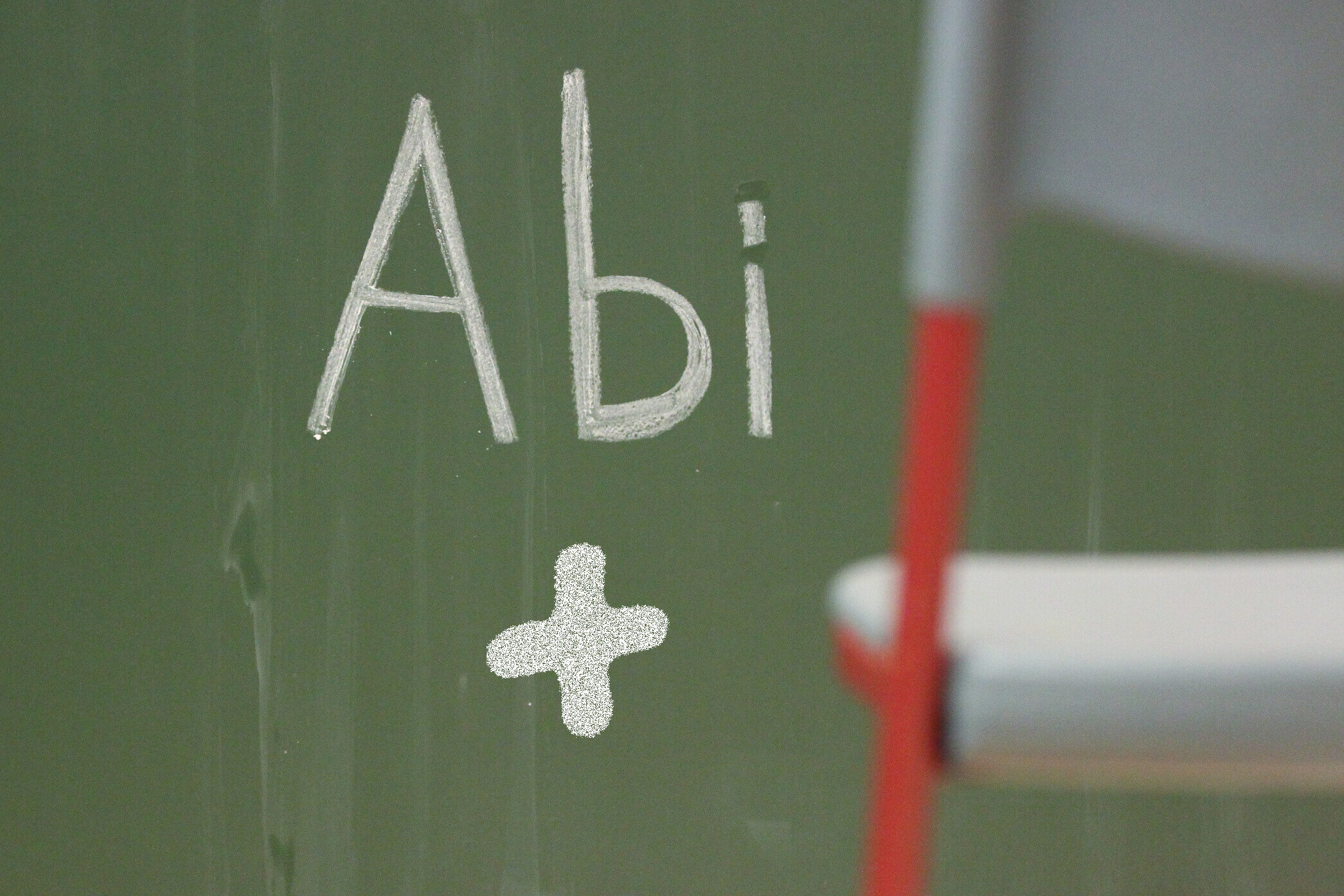 "Abi" und ein Pluszeichen auf grüner Tafel. Stuhl rechts unscharf.