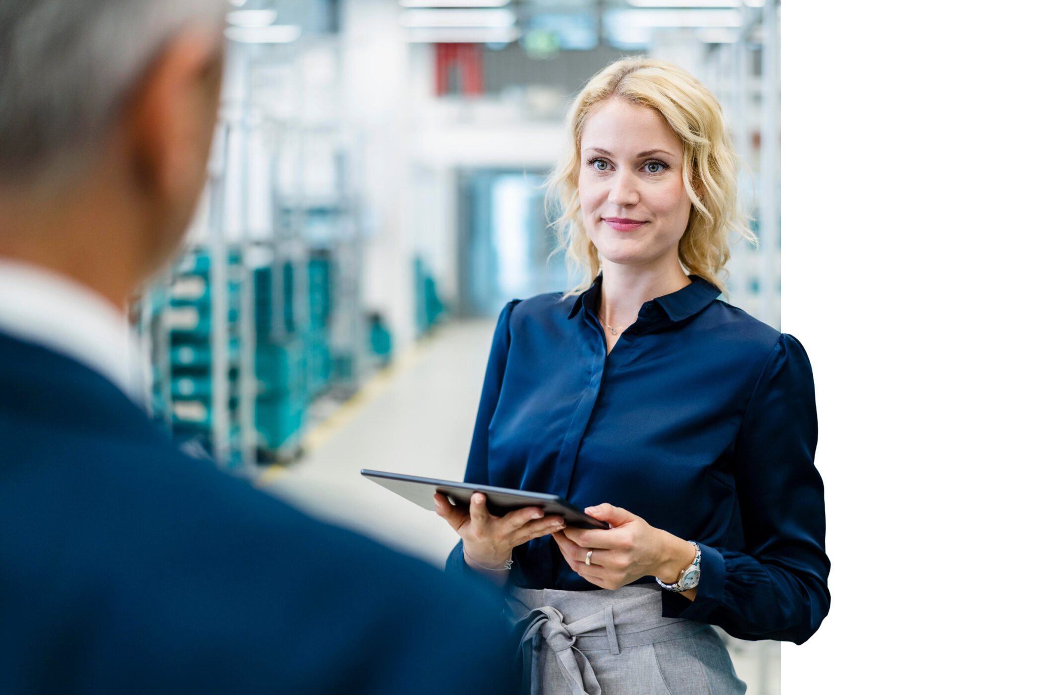Frau mit Tablet in blauem Hemd, steht in einem Büro. Eine Person im Vordergrund.
