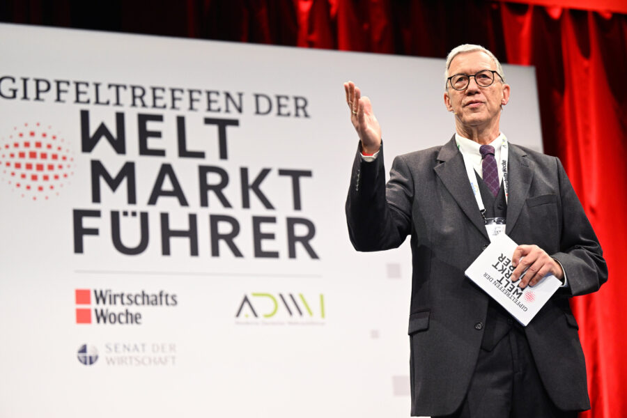 Mann auf einer Bühne, spricht, hält Notizblock, Hintergrundtext: "Gipfeltreffen der Weltmarktführer".