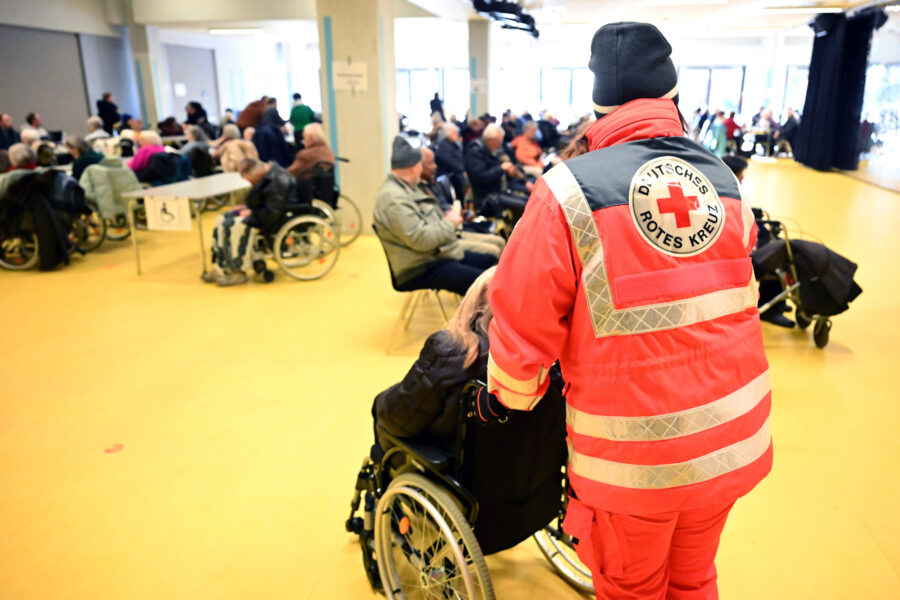 Viele Menschen in einem Raum, Rollstuhl, Person in rotem Rotkreuz-Anzug.