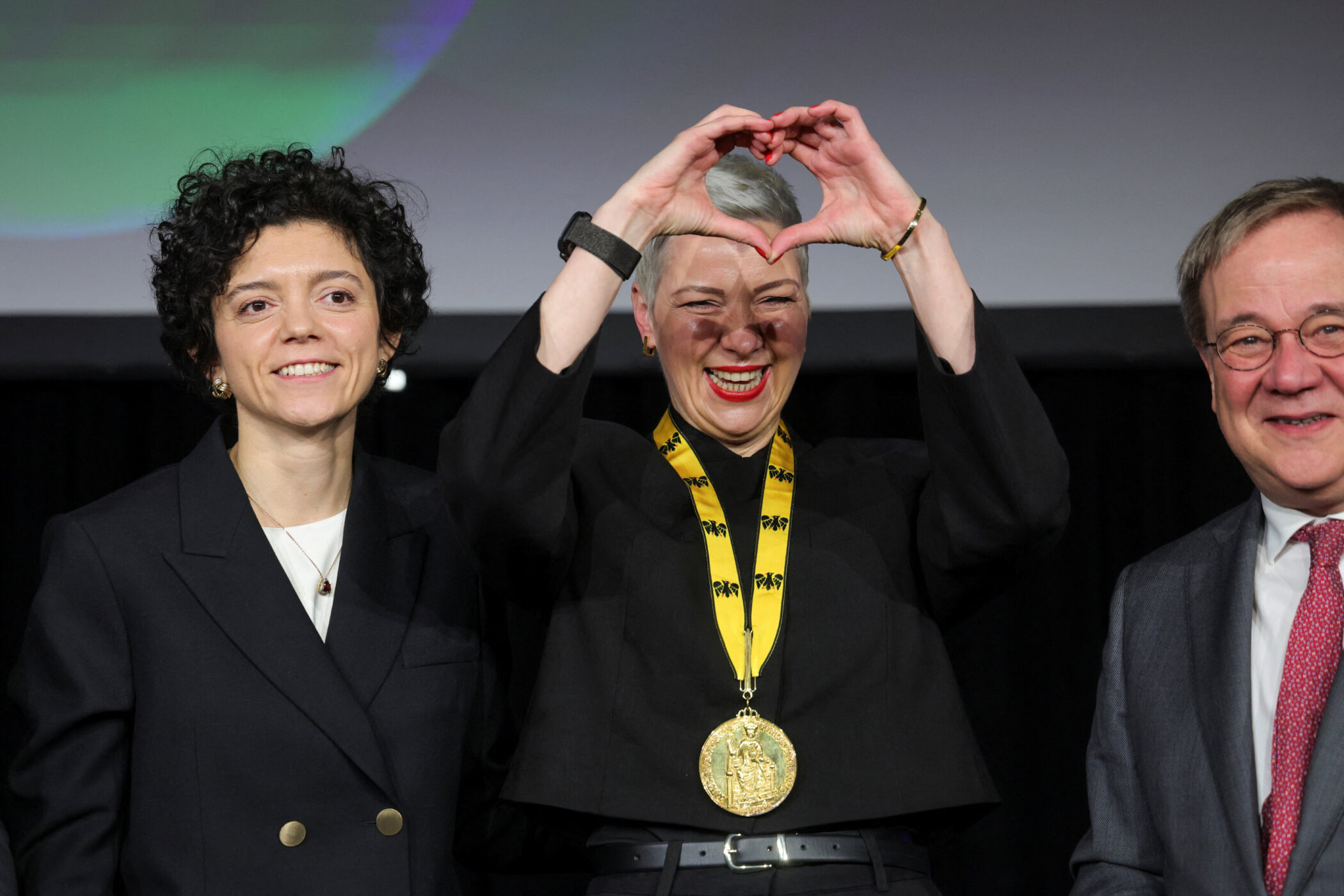Drei Personen stehen zusammen, die mittlere Person formt ein Herz mit den Händen.