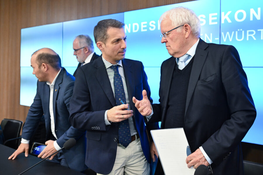 Vier Männer in Anzügen, zwei im Gespräch, einer hält ein Tablet.