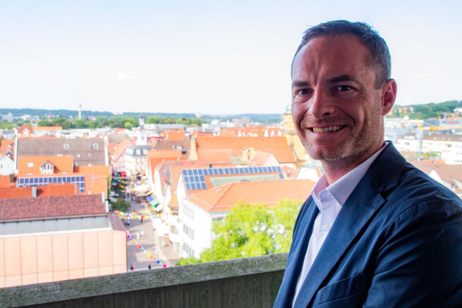 Mann in Anzug lächelt, im Hintergrund Stadtansicht mit roten Dächern.