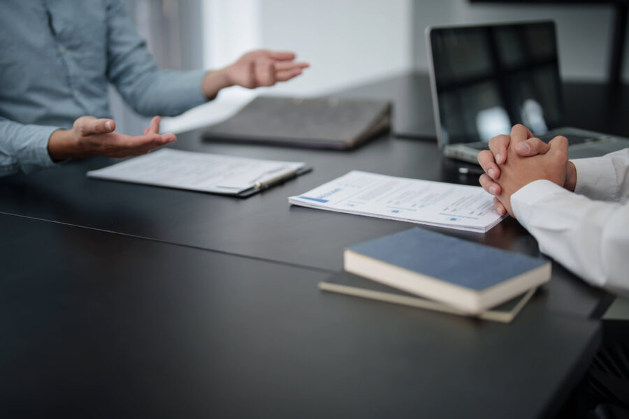 Zwei Personen an einem Tisch mit Papieren und Laptop. Hände gestikulieren und gefaltet.