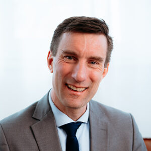 Mann in grauem Anzug und dunkler Krawatte lächelt in die Kamera vor hellem Hintergrund.