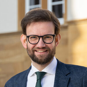 Person mit Brille und Bart lächelt im Anzug vor einem Gebäude.