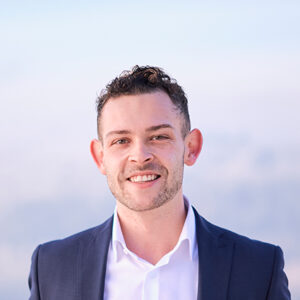 Ein lächelnder Mann im Anzug vor unscharfem Hintergrund.