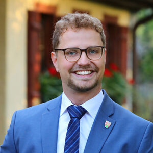 Mann mit Brille im blauen Anzug und Krawatte, lächelt vor verschwommenem Hintergrund.