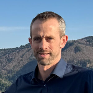 Person vor bewaldeter Berglandschaft, trägt ein blaues Hemd.