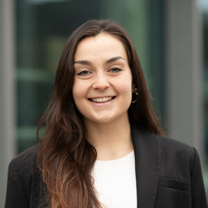 Junge Frau mit langen Haaren, schwarzem Blazer vor unscharfem Hintergrund.