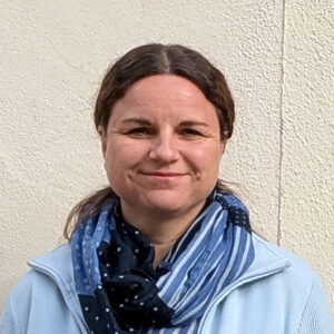 Eine Person in blauer Jacke und Schal vor einer weißen Wand.