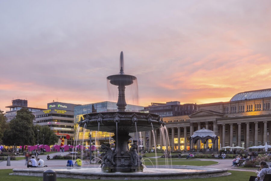 Springbrunnen im Park bei Sonnenuntergang, Menschen sitzen auf Bänken, Gebäude im Hintergrund.