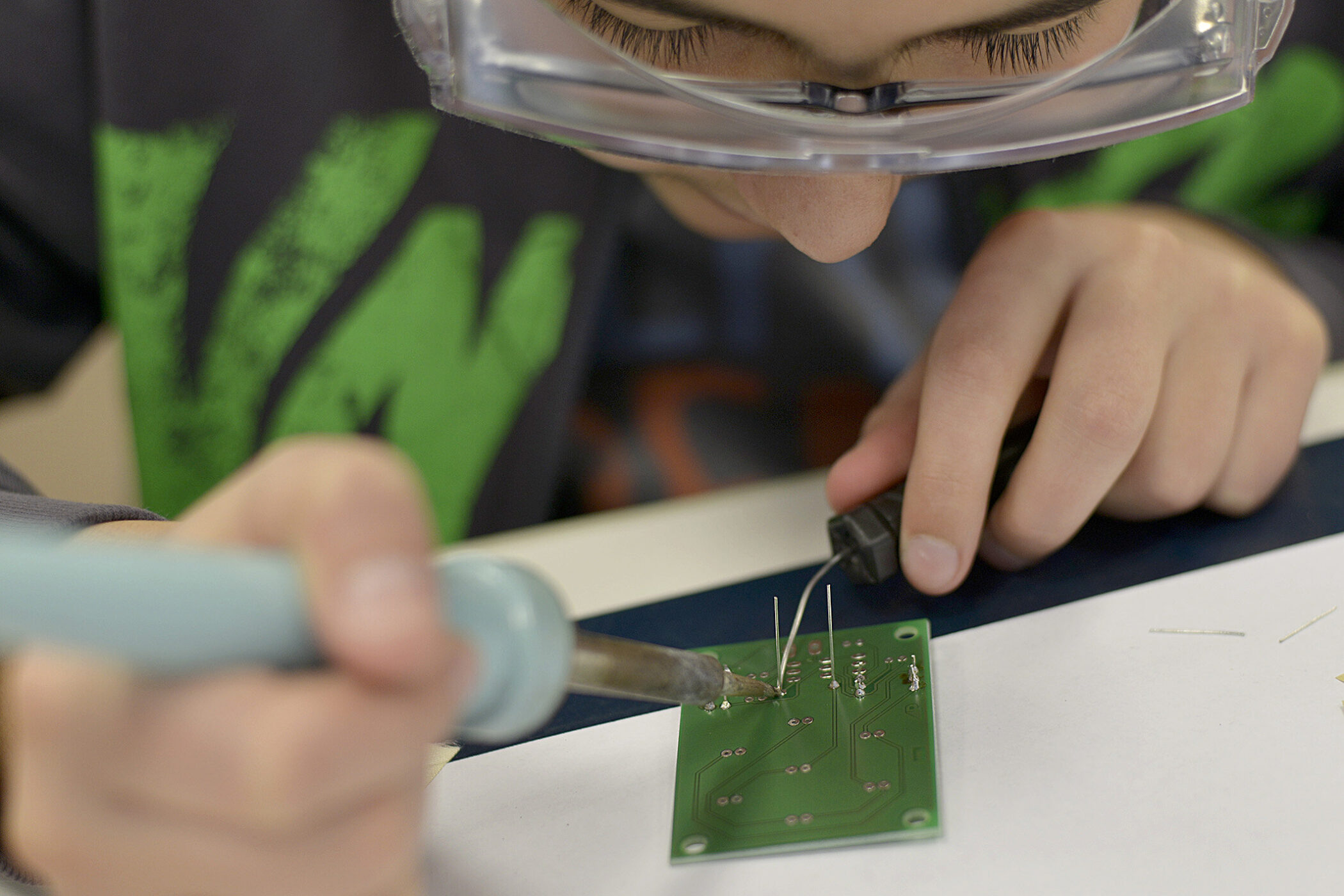 Person lötet an grüner Leiterplatte, trägt Schutzbrille.