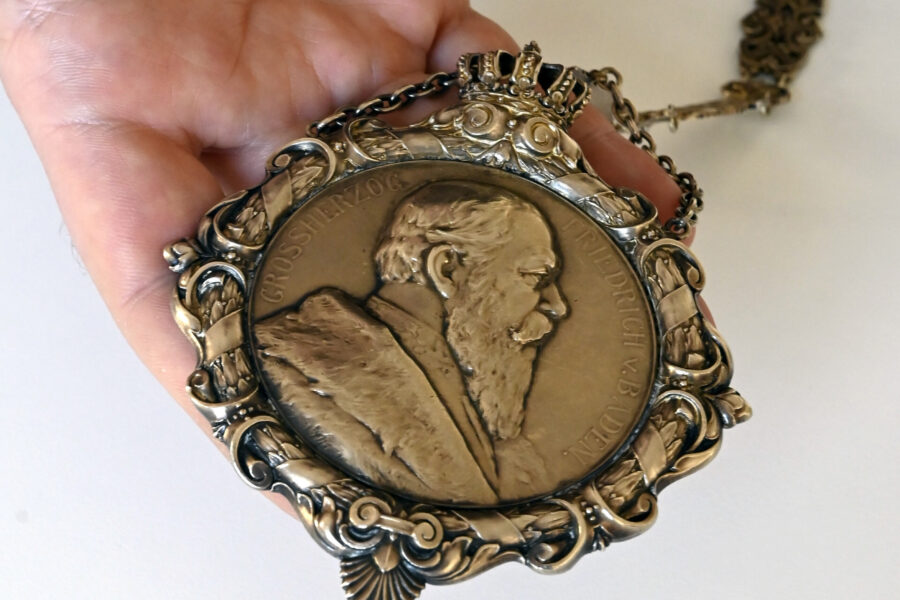 Hand hält metallene Medaille mit Relief eines bärtigen Mannes, umrahmt von Verzierungen.