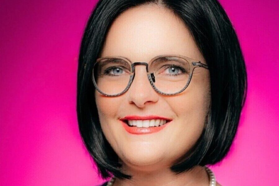 Frau mit Brille und kurzen dunklen Haaren vor pinkem Hintergrund.