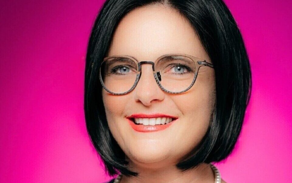 Frau mit Brille und kurzen dunklen Haaren vor pinkem Hintergrund.