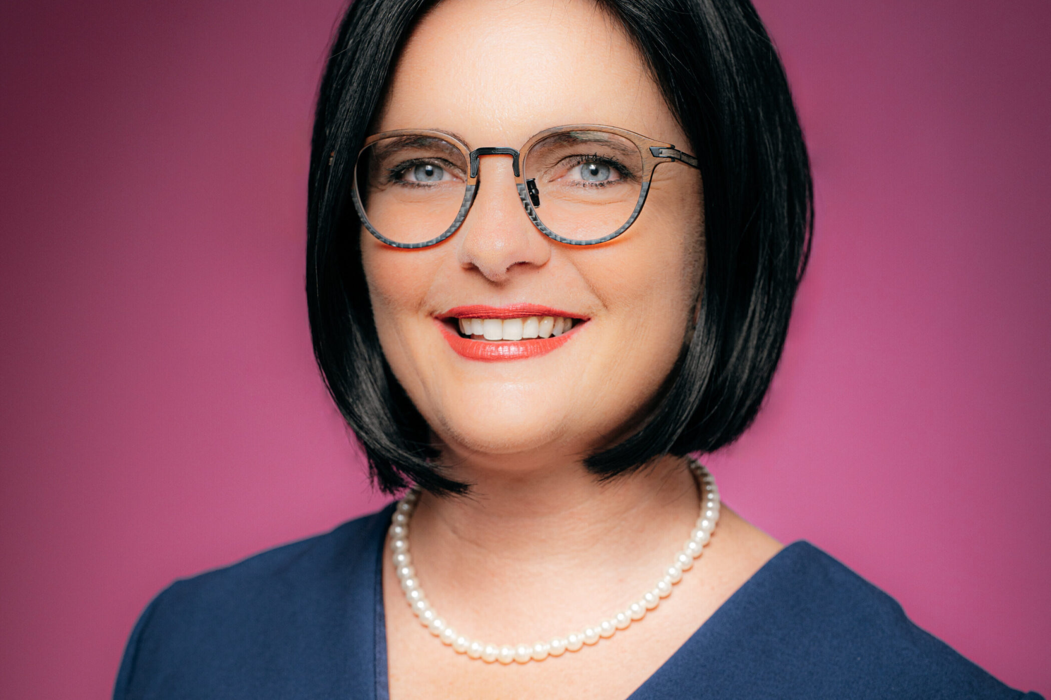 Frau mit Brille, schwarzem Haar, Perlenkette vor pinkem Hintergrund.