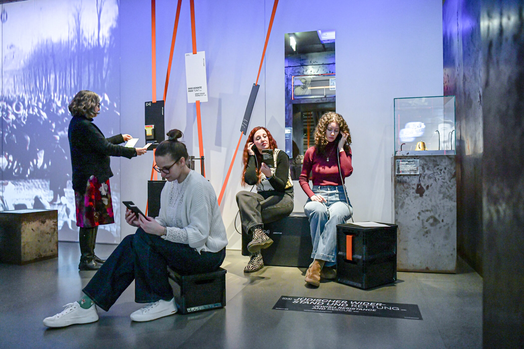 Vier Personen in modernem Ausstellungsraum, teilweise mit Kopfhörern und Smartphones.