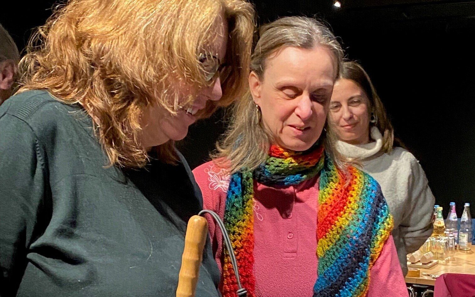 Drei Personen, zwei vorn, eine im Hintergrund. Tisch mit Flaschen und Gläsern dahinter.