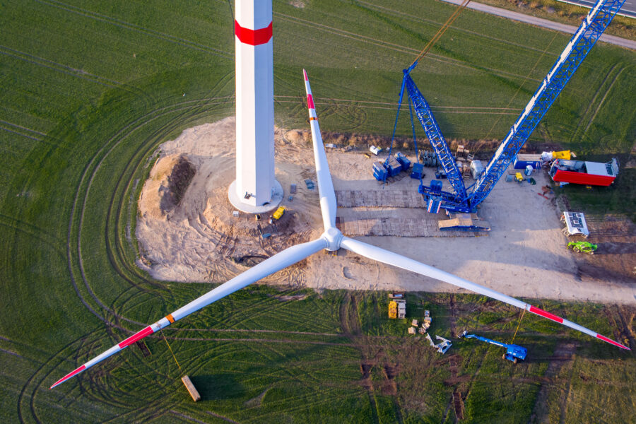 Windrad im Bau, großer Kran, grüne Wiese, Betonsockel.