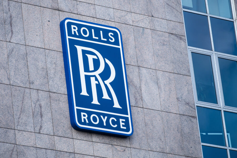 Blaues Rolls-Royce-Logo an grauer Gebäudewand mit Fenstern rechts.