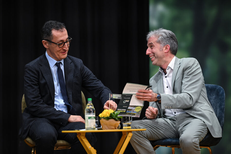 Zwei Männer im Anzug sitzen, einer hält ein Papier, Tisch mit gelber Blume und Wasserflasche.