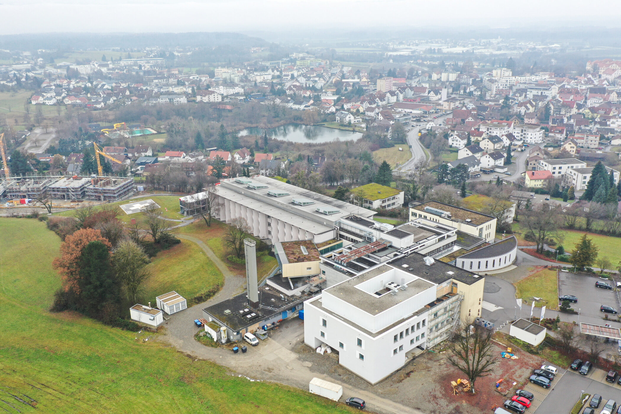 Gebäude mit Flachdach und Vorplatz, umgeben von Wiesen, Bäumen und Wohngebäuden.