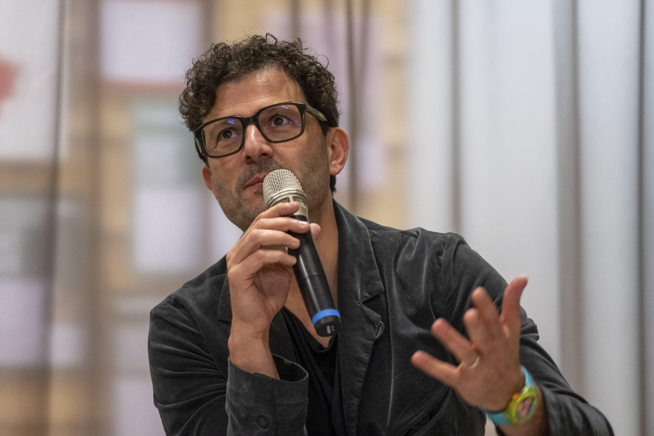 Mann mit Brille spricht in ein Mikrofon, Gestik mit der linken Hand.