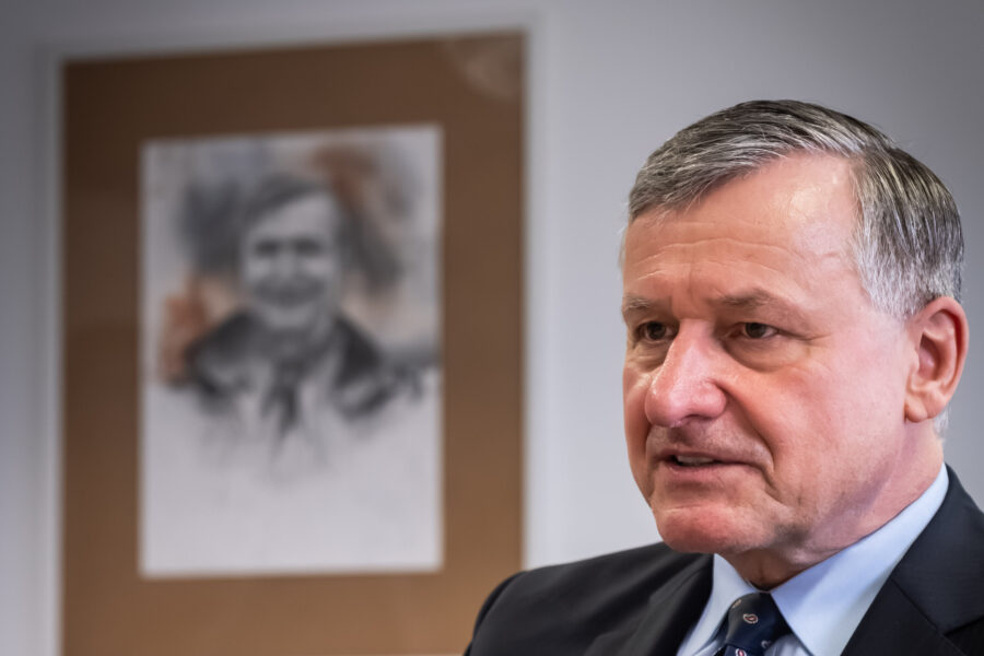 Älterer Mann in Anzug vor unscharfem Porträt im Hintergrund.