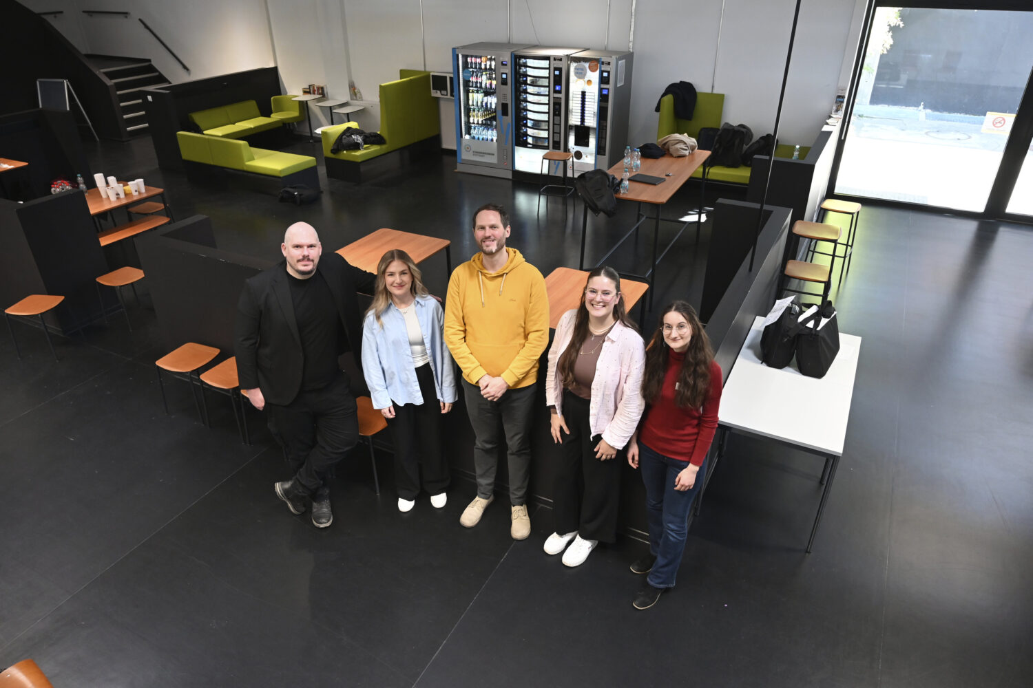 Fünf Personen stehen in einem Raum mit Tischen und Sitzgelegenheiten.