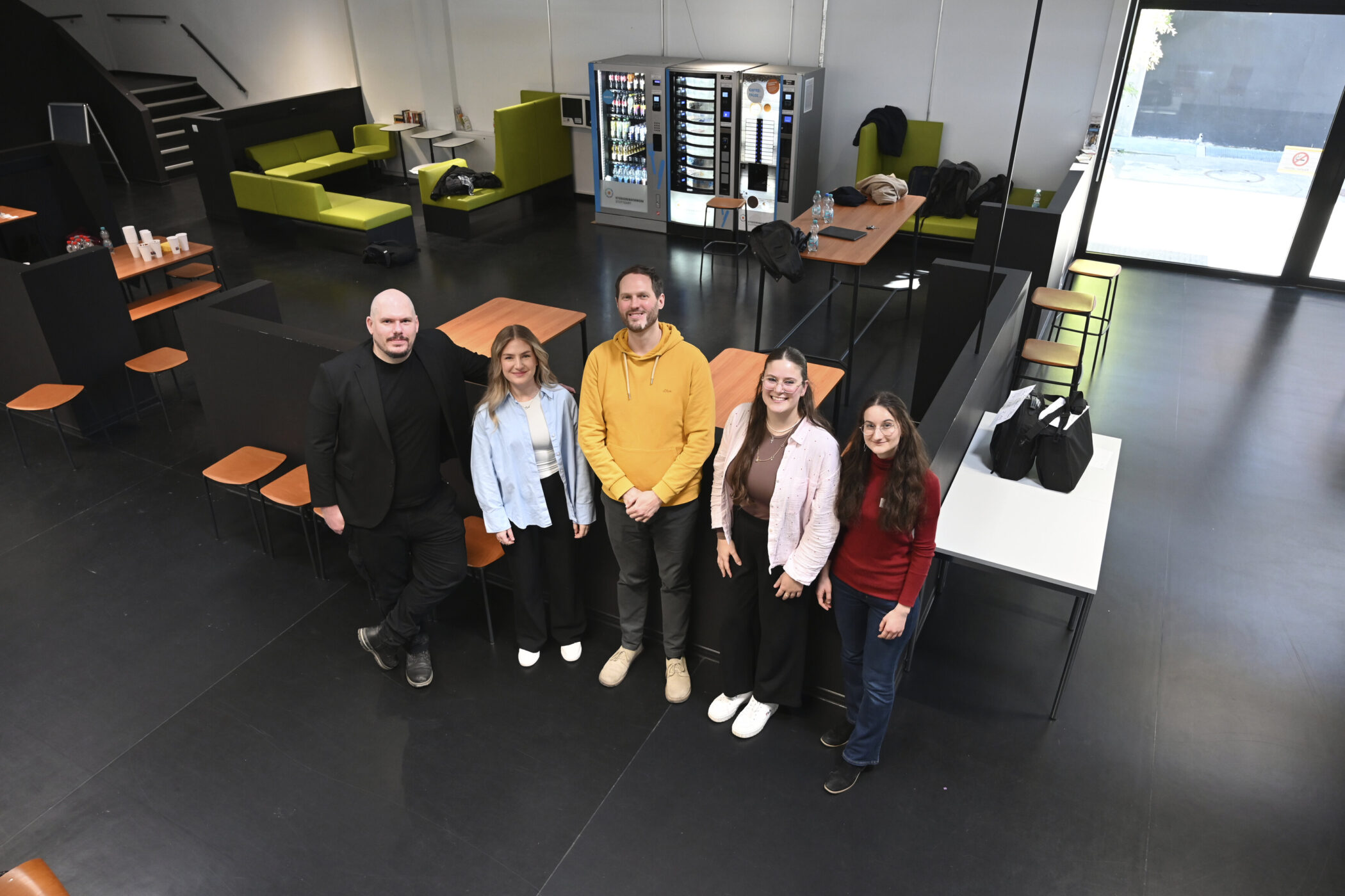 Fünf Personen stehen in einem Raum mit Tischen und Sitzgelegenheiten.