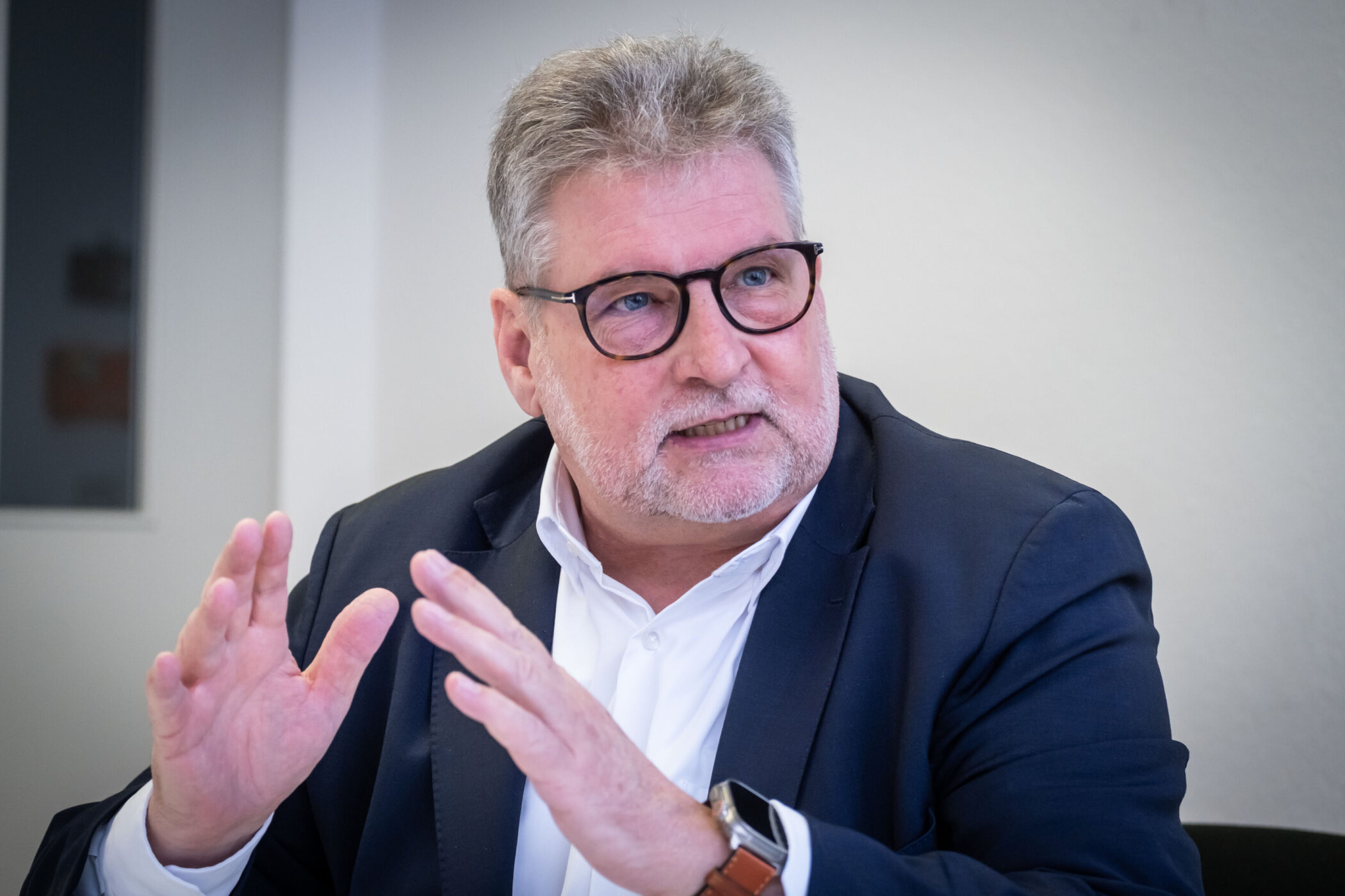 Älterer Mann mit Brille gestikuliert, trägt Anzug und Hemd.