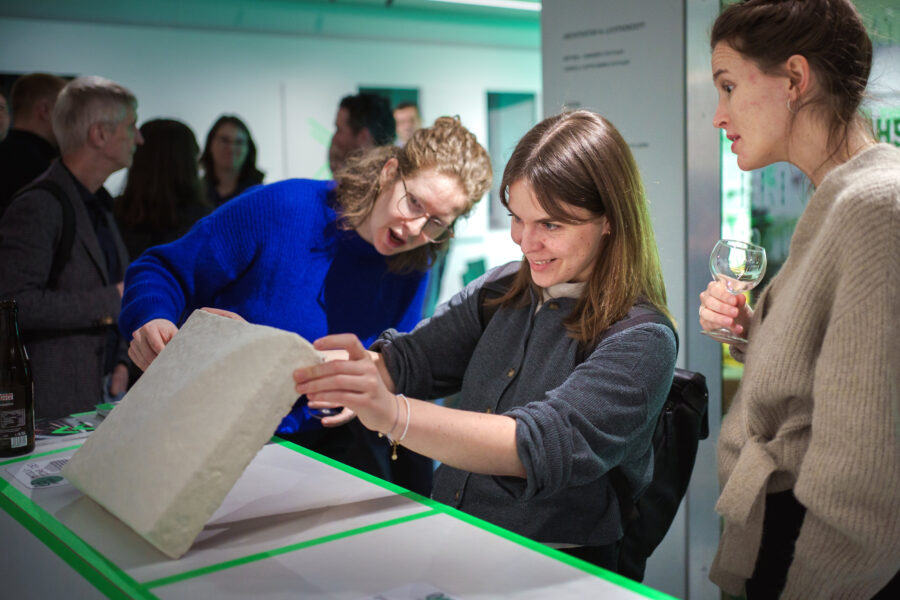 Drei Frauen betrachten ein Objekt auf einem Tisch, eine hält ein Weinglas.