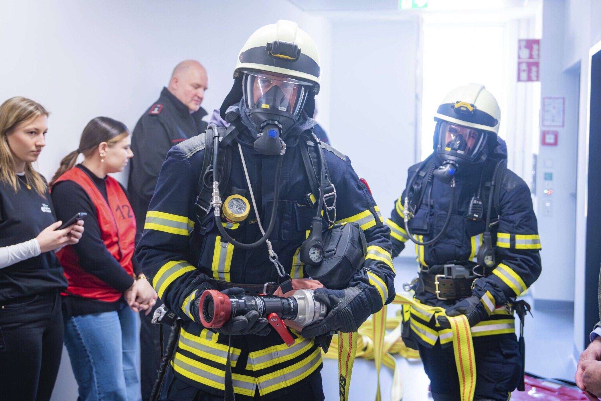 Zwei Feuerwehrleute in Uniformen mit Atemgeräten, umgeben von mehreren Personen.