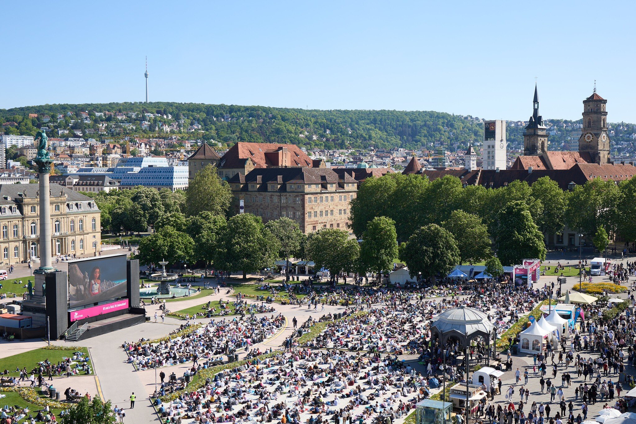 Viele Menschen auf einem Platz mit Leinwand, historischen Gebäuden und Hügel im Hintergrund.