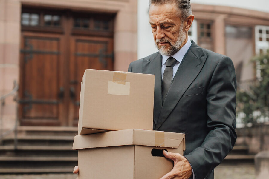 Mann in Anzug trägt zwei Kartons vor einem Gebäude mit Holztüren.