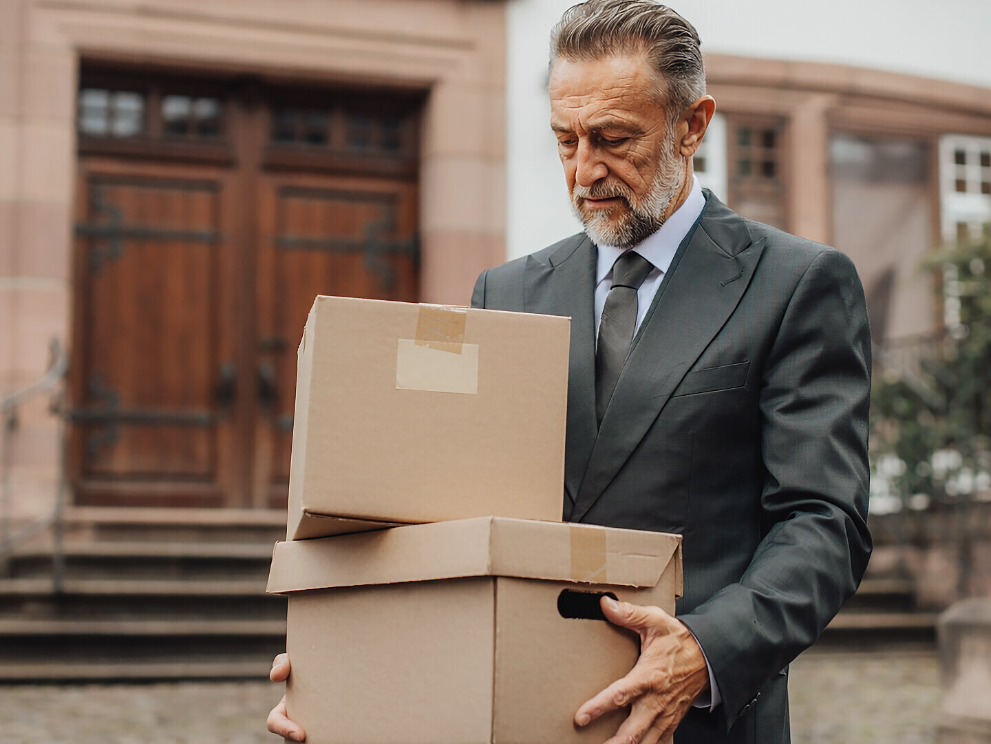 Mann in Anzug trägt zwei Kartons vor einem Gebäude mit Holztüren.