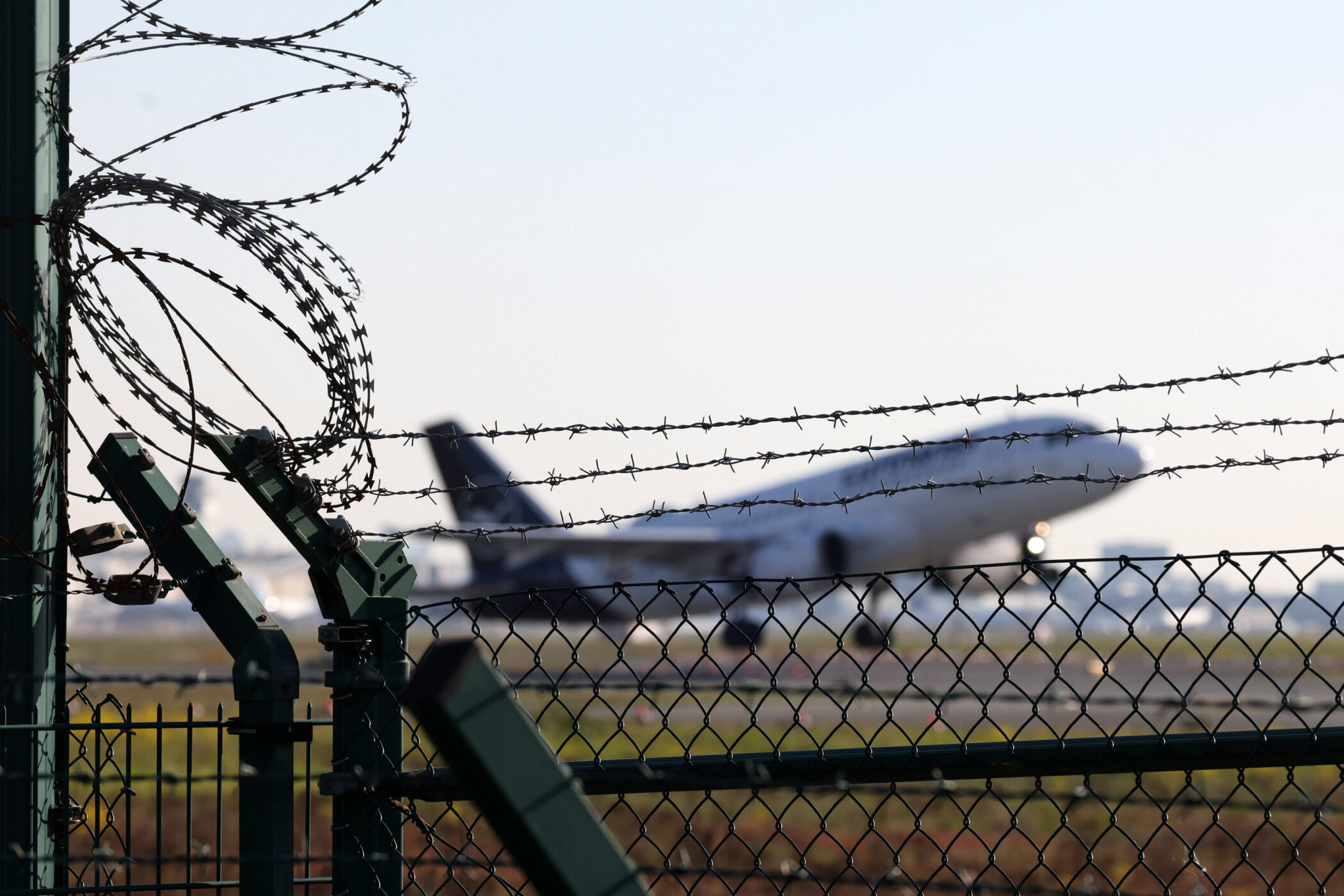 Zaun mit Stacheldraht, unscharfes Flugzeug im Hintergrund.