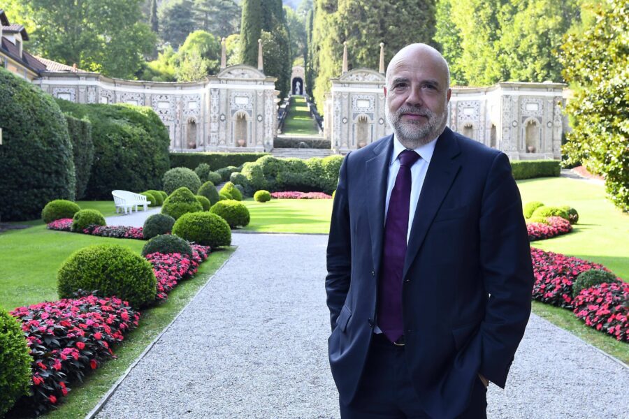 Ein Mann im Anzug steht lächelnd in einem Garten mit gepflegten Hecken und Blumen.