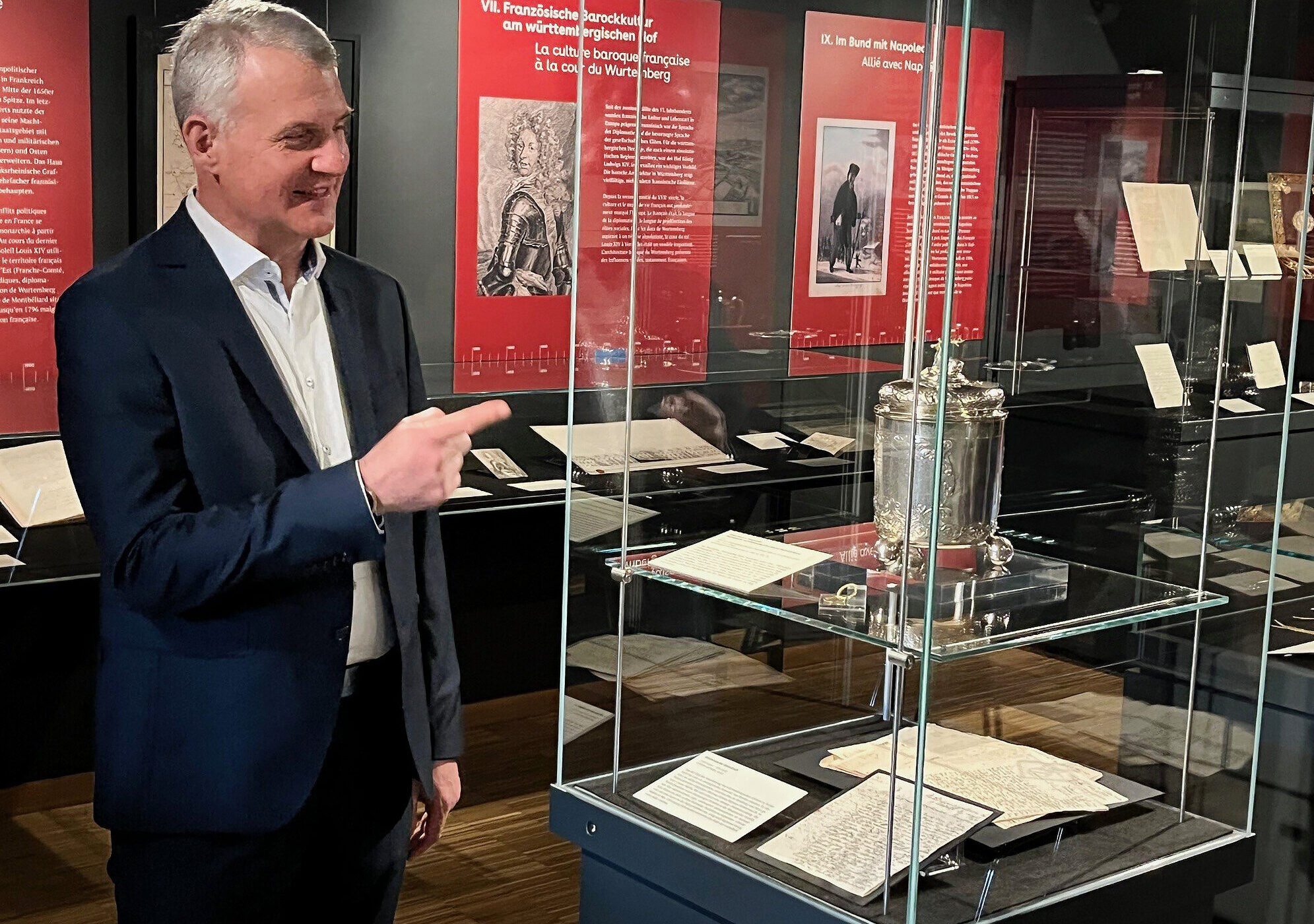 Ein Mann im Anzug zeigt in einem Museum auf eine Glasvitrine mit Exponaten.